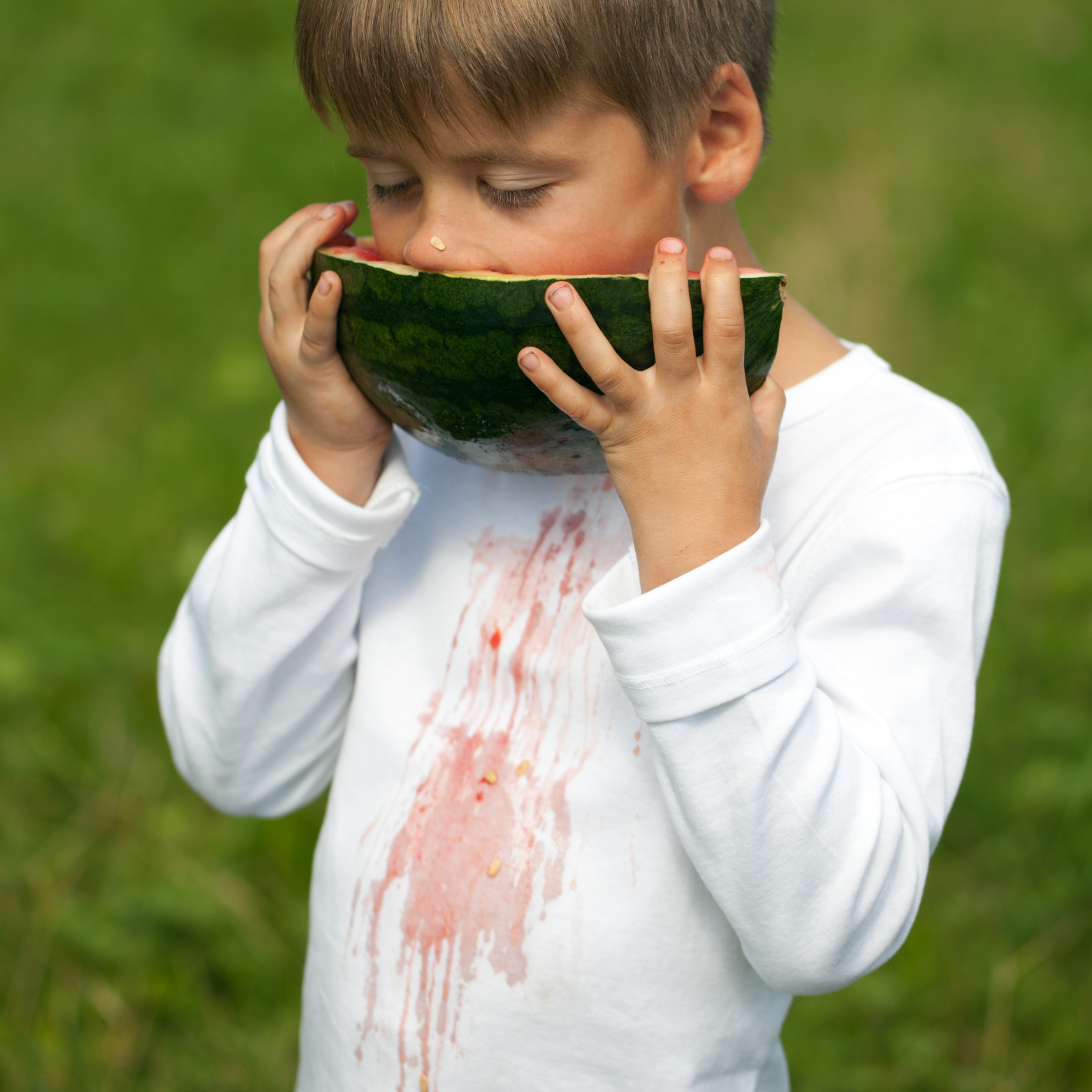 Photo d’un jeune garçon mangeant une tranche de pastèque, avec des taches de jus rouge sur son t-shirt blanc, illustrant une scène d’été naturelle et joyeuse.
